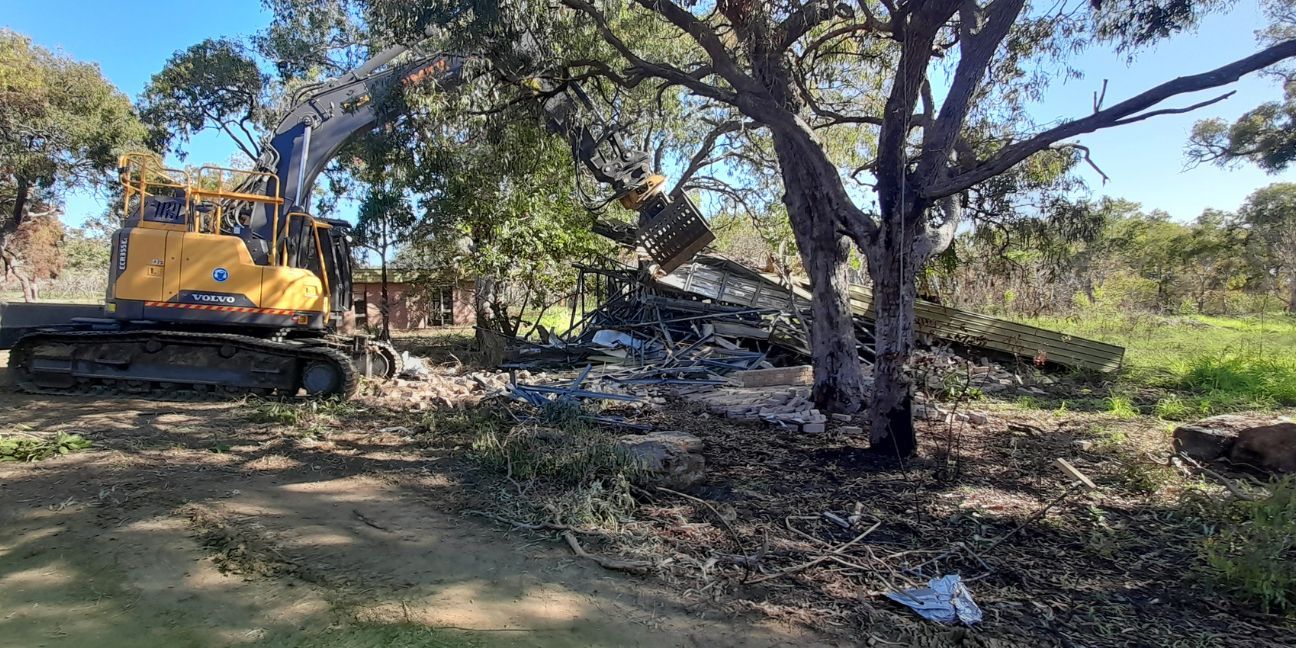 Foreground shows a demolished villa and demolition machinery, with a still-standing villa in the background.