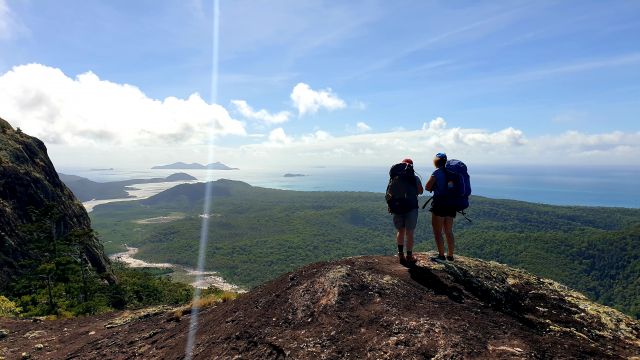Two people wearing large backpacks standing atop of a large rock looking at a view across an island towards the ocean and other islands. 