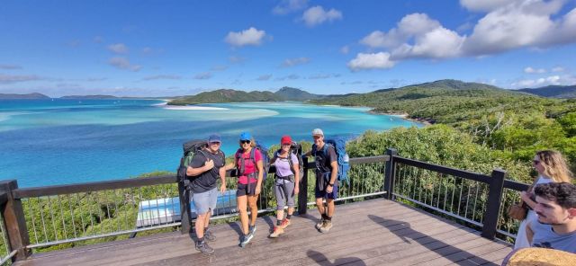 Four people wearing large backpacks standing on a timber lookout with views of turquoise ocean and green island in the background. 