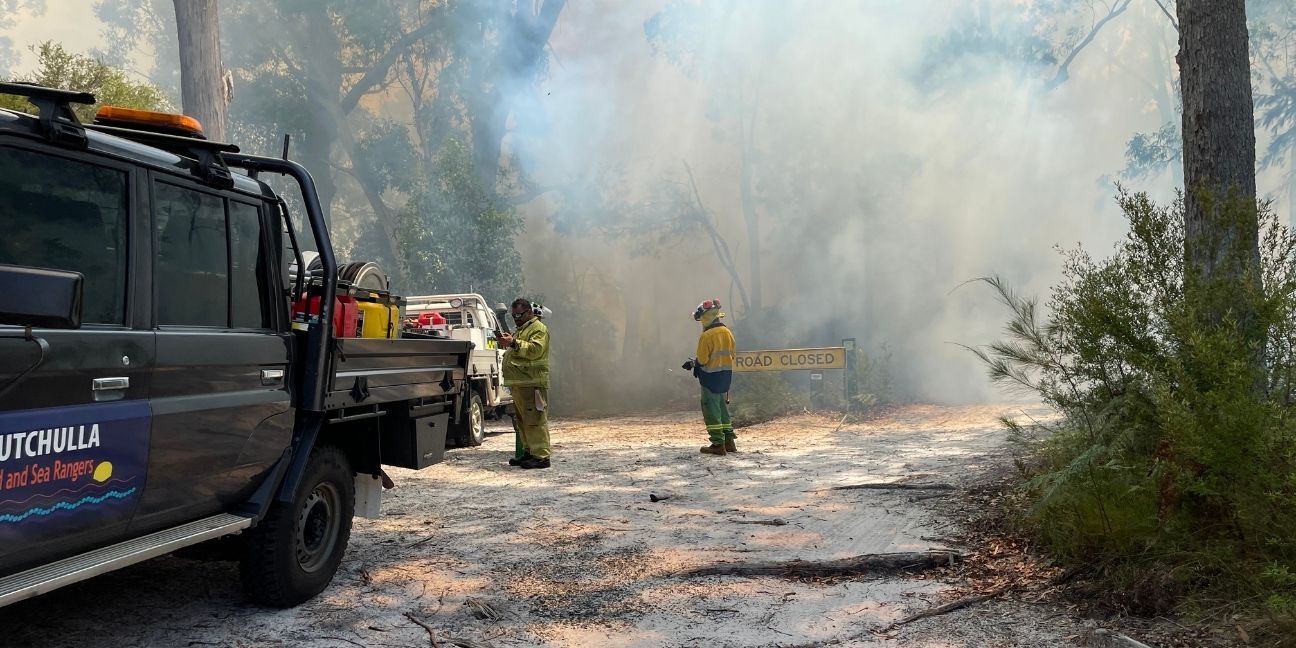 Two people in fire gear standing in front of a road closed sign with smoke behind it. There's also a couple of vehicles on the left-hand side.