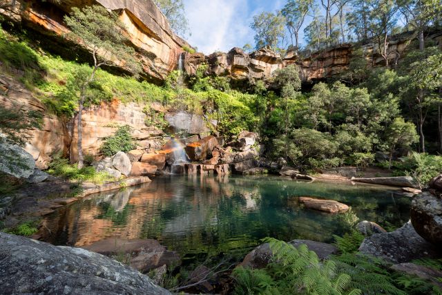 A serene natural rock pool located at Gudda Gumoo Gorge. The rock has clear water surrounded by rocky cliffs and lush green vegetation under a partly cloudy sky, the water of the pool reflects the surrounding trees and rocks.
