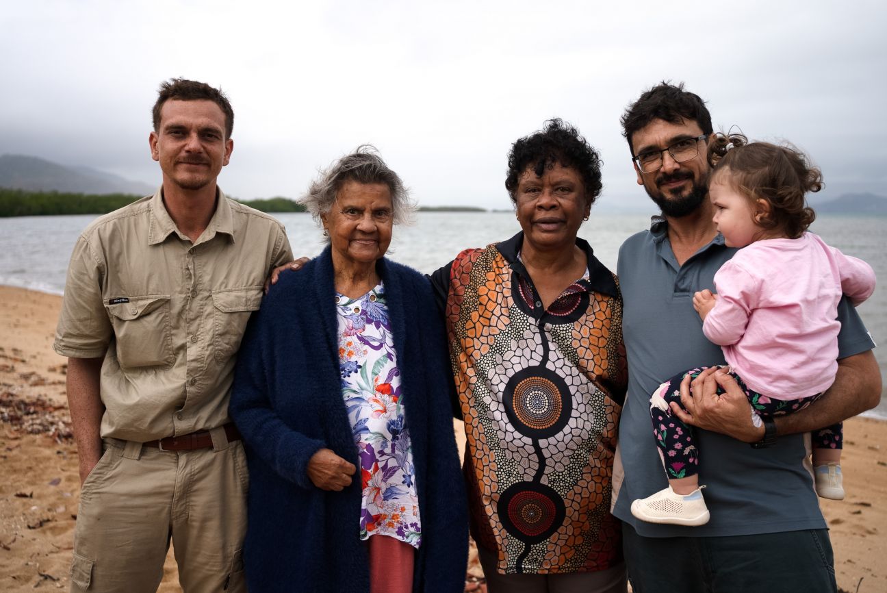 Five people standing on a beach with a stormy sky in the background. Two Wulgurukaba female Edlers in the middle with two Wulgurukaba younger men on either side, one man is holding a young child. 