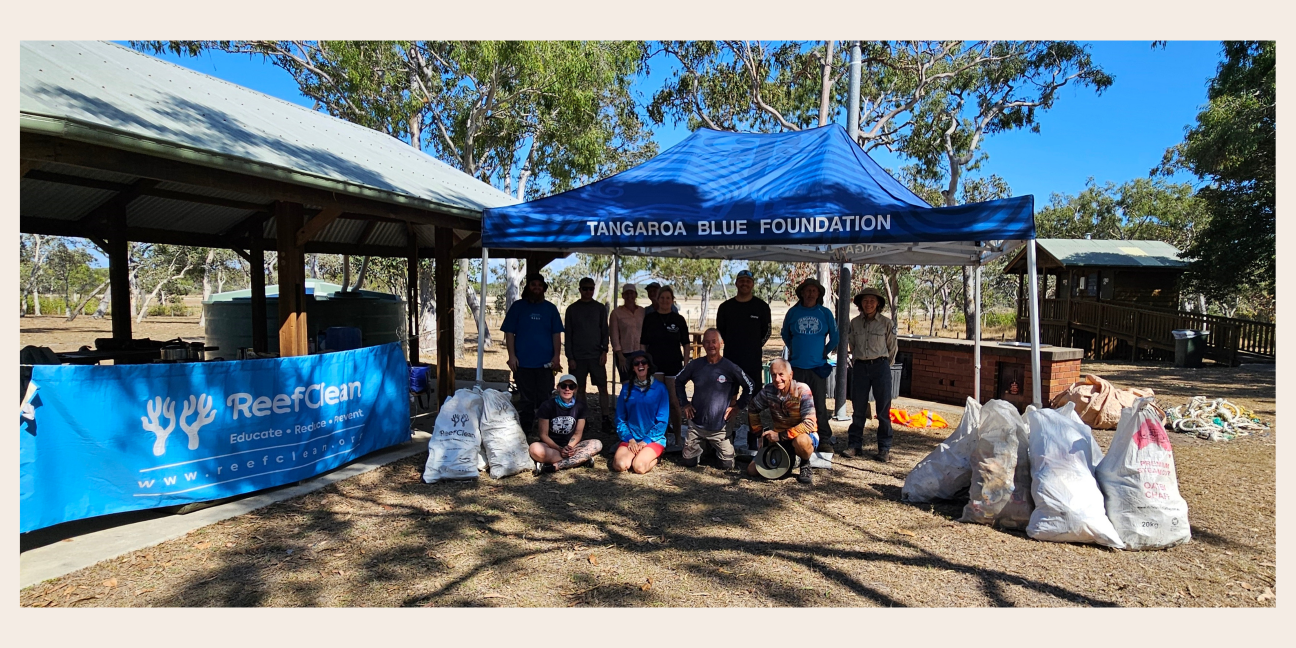 A group of volunteers, Rangers and members of the Tangaroa Blue Foundation congregated under a blue gazebo labelled Tangaroa Blue Foundation. The people are surrounded by large bags of collected rubbish and have a Reef Clean banner hanging up to their left, attached to a picnic shelter.