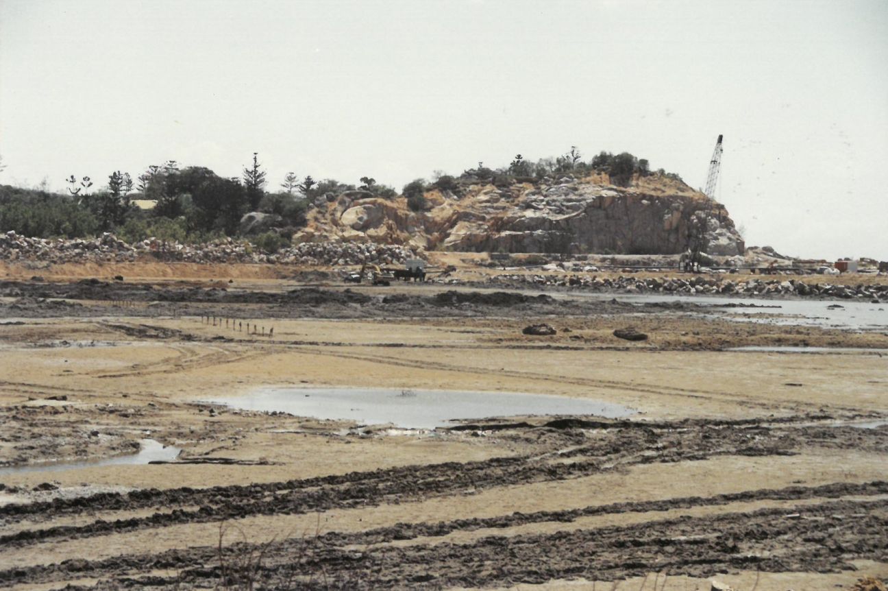 Construction site with sand and mud in foreground, cranes, rock wall and destroyed headland in background. 