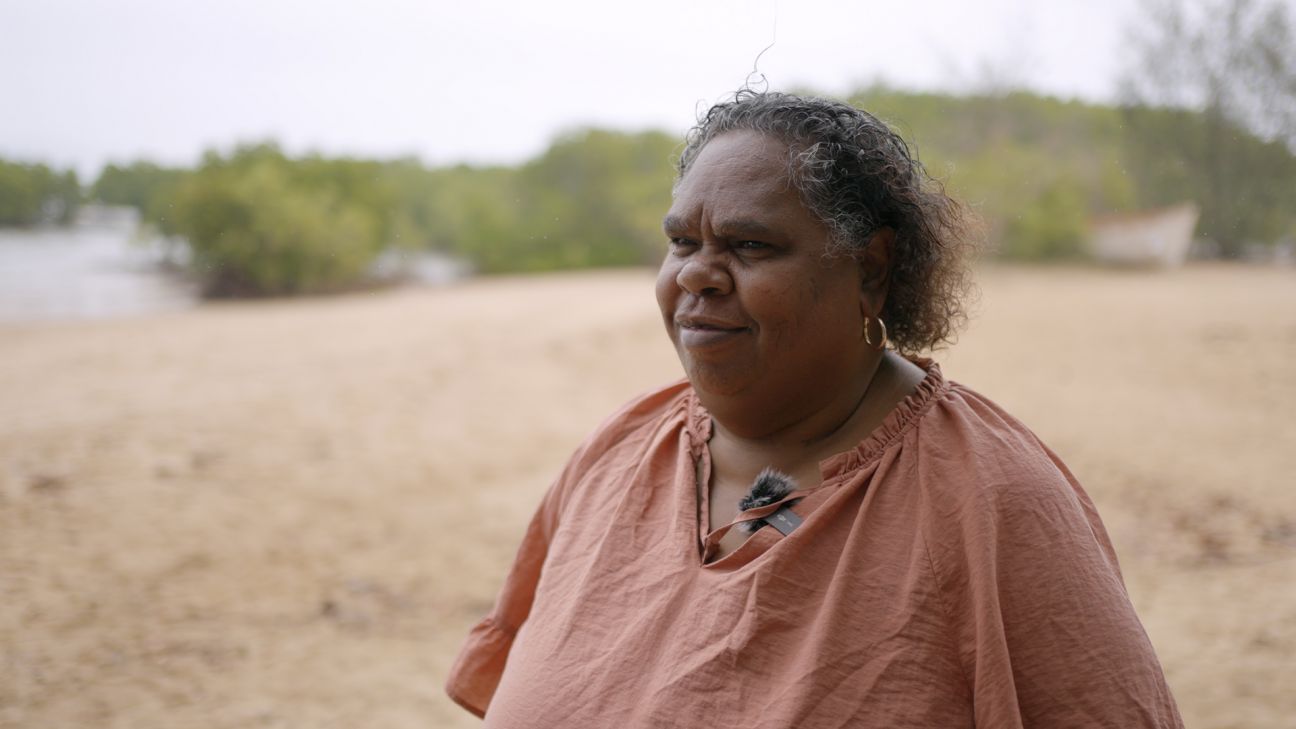 Wulgurukaba female Elder standing on a beach with mangrove trees in background.  