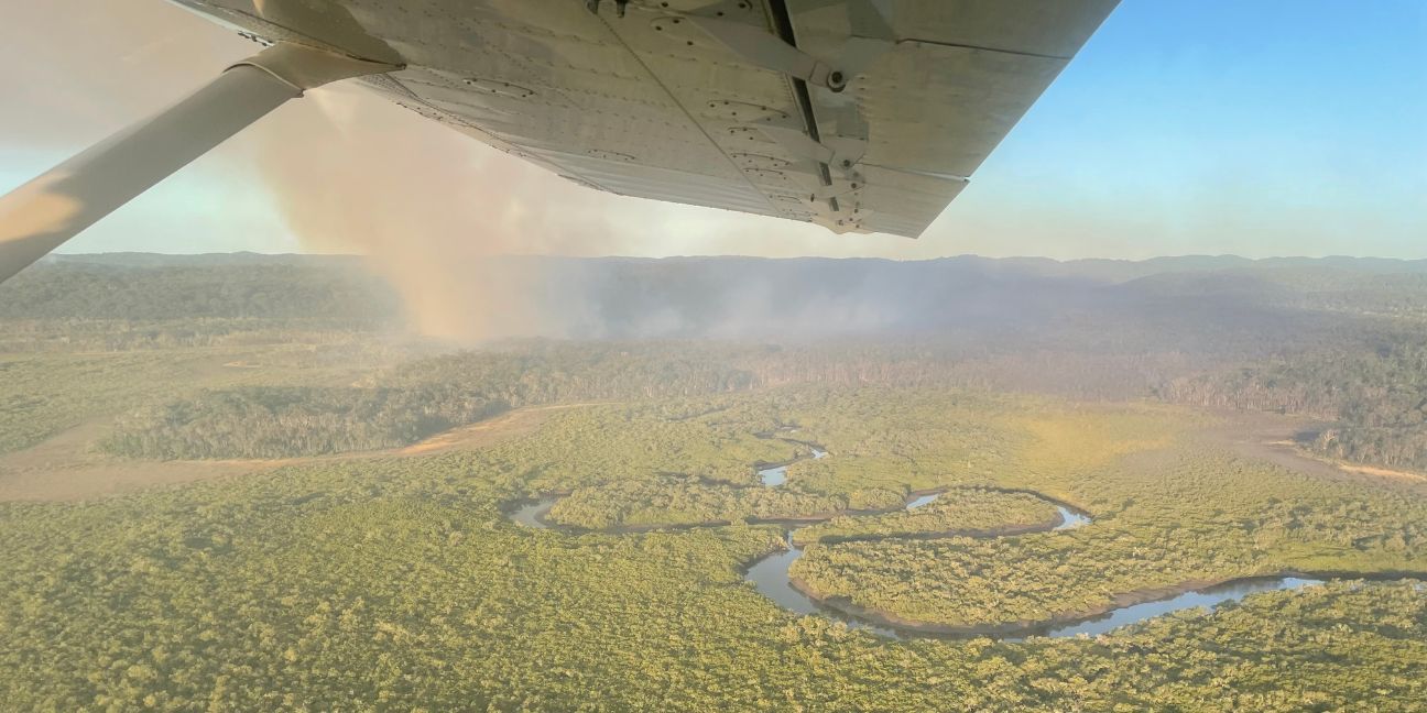 An ariel view of a planned burn on K’gari. Brown smoke rises from the rainforest, nearby a winding creek.  