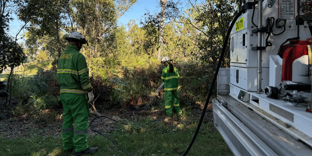 Two Rangers in green fire uniforms and white helmets use a fire hose from their vehicle to put out a test burn.