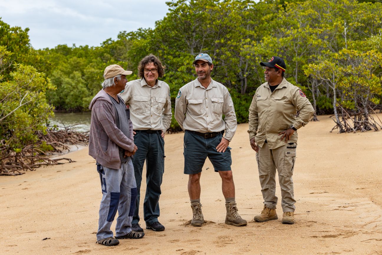 Four men standing on a beach with mangroves in the background. Two Wulgurukaba men, one wearing plain clothes, other man wearing uniform. Two men wearing Ranger uniforms.