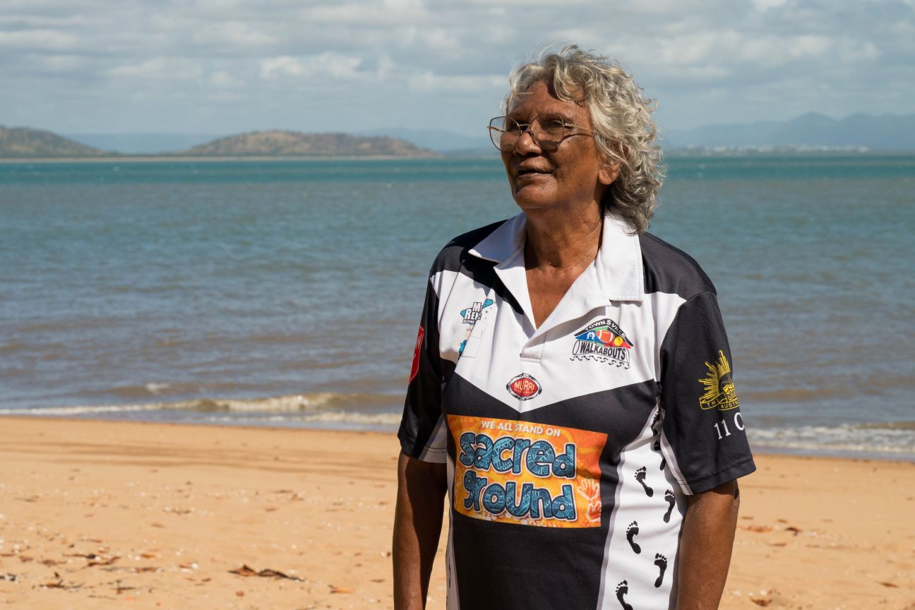 Wulgurukaba female elder standing with hands on hips on a beach with mainland in background.