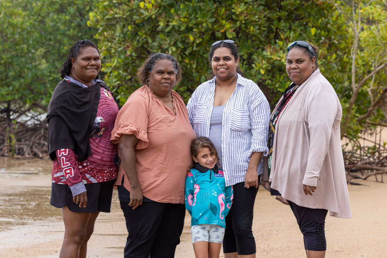 Four Wulgurukaba women standing side by side with a young girl in front on a beach with mangroves in background. 