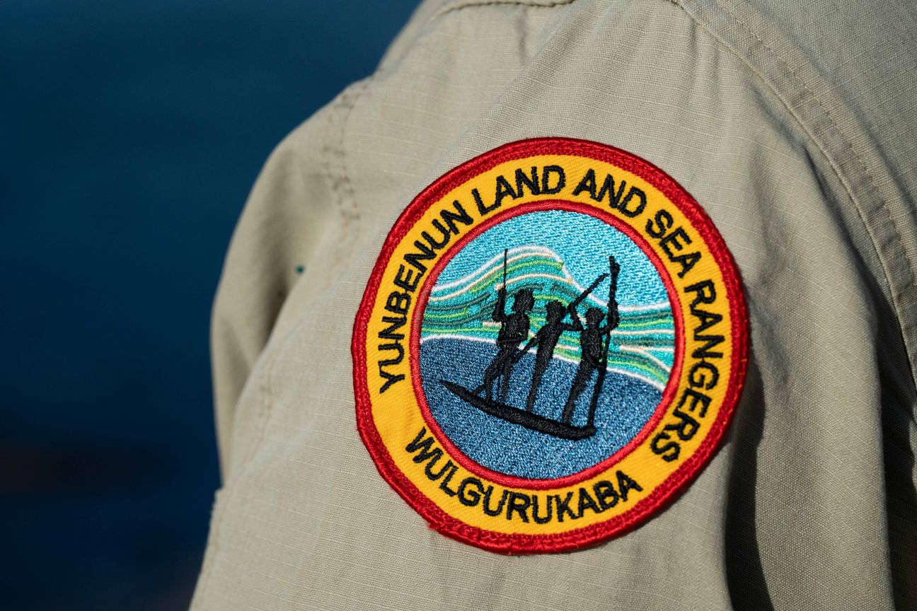 Badge on should of shirt of Ranger uniform. Three black people standing in the canoe with paddles on blue and green background in centre of badge. Wulgurukaba Yunbenun Land and Sea Rangers words written in black with yellow background with red outline around outside of badge.   