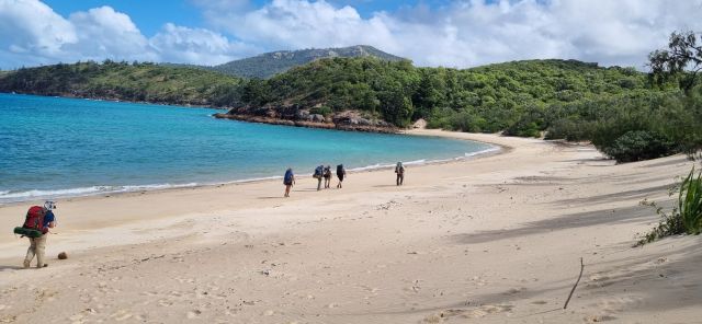 A group of people wearing parge backpacks walking on a beach. 