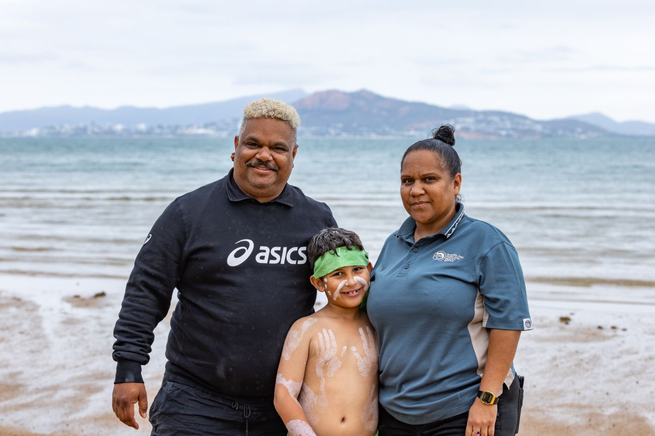 Three Wulgurukaba people standing on a beach with Townsville in the background. Male and female with male child dressed as an aboriginal dancer. 