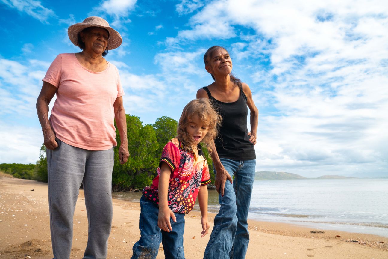 Three Wulgurukaba people standing on a beach with mangroves, ocean and blue sky in the background. Older woman, younger woman and young boy.