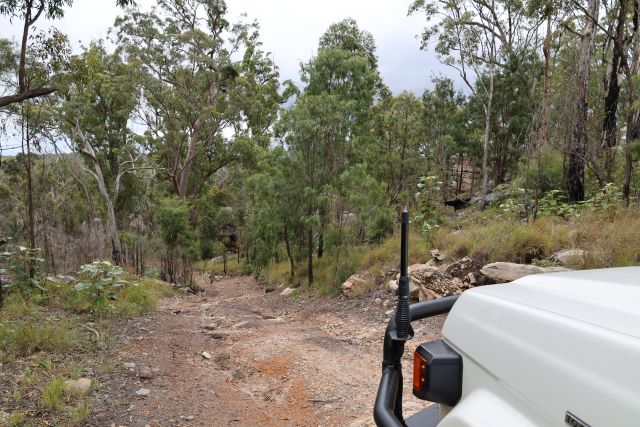 A rugged off-road vehicle track showing a rocky dirt track surrounded by dense trees and vegetation. A portion of a white 4WD vehicle is visible on the right.