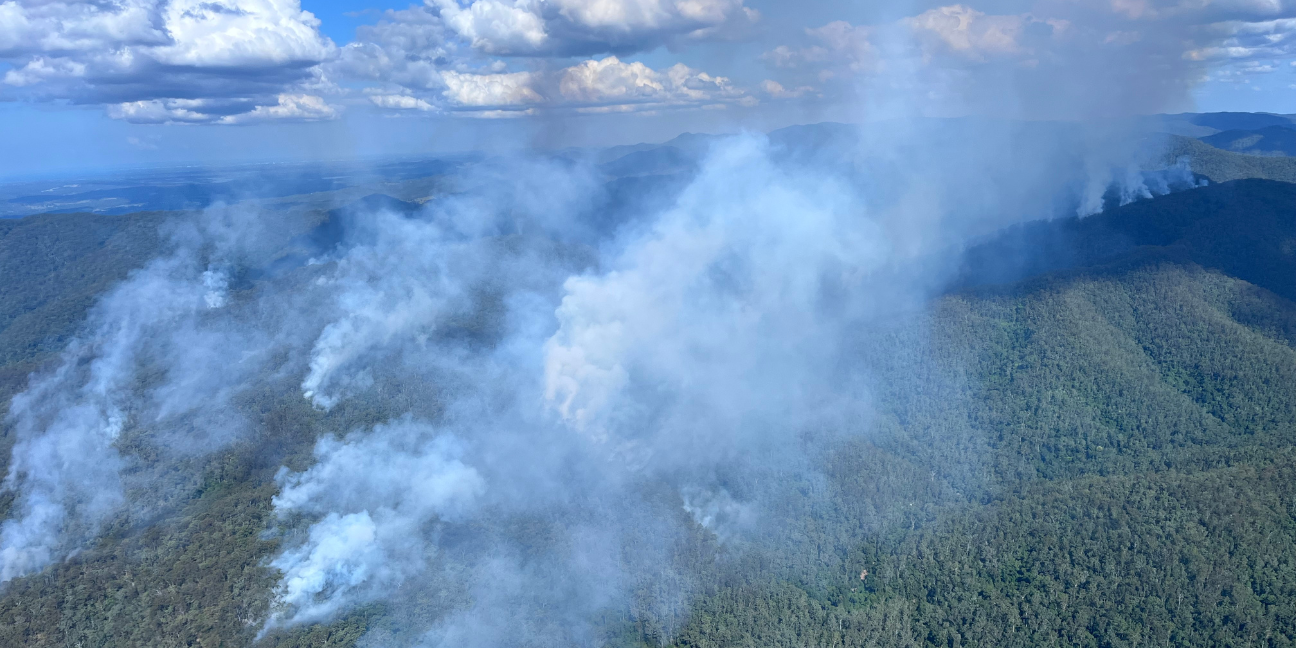 An aerial view of smoke emitting from dense bushland across mountains