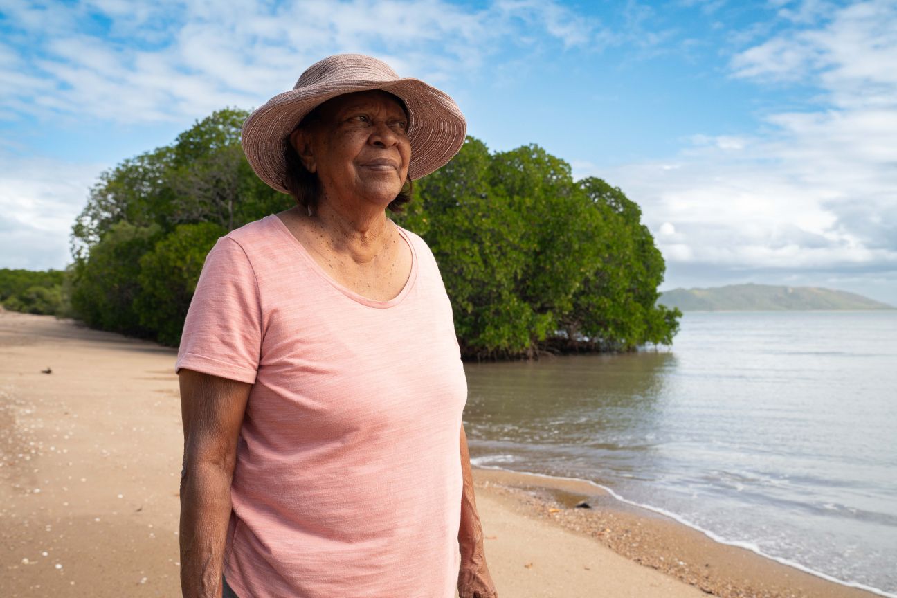 Wulgurukaba female Elder standing on a beach with ocean and mangroves in the background. 