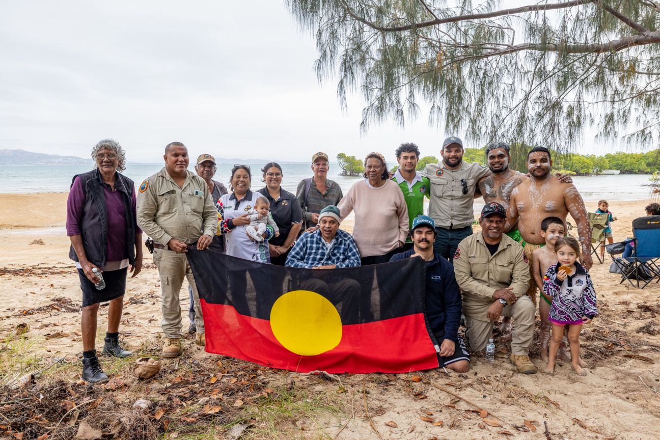Large group of Wulgurukaba people of various ages standing and kneeling on a beach in front of a black, red and yellow Aboriginal flag.  