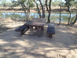 A wooden picnic table with two wooden benches situated on sandy ground near Nowranie Waterhole, surrounded by trees.