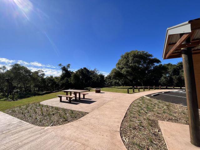 A concrete pathway leads to a wheelchair-accessible picnic table and firepit on the left and car parking on the right. Behind the pathway is a sloped grassy area with a shaded picnic table and firepits, surrounded by a background of trees.