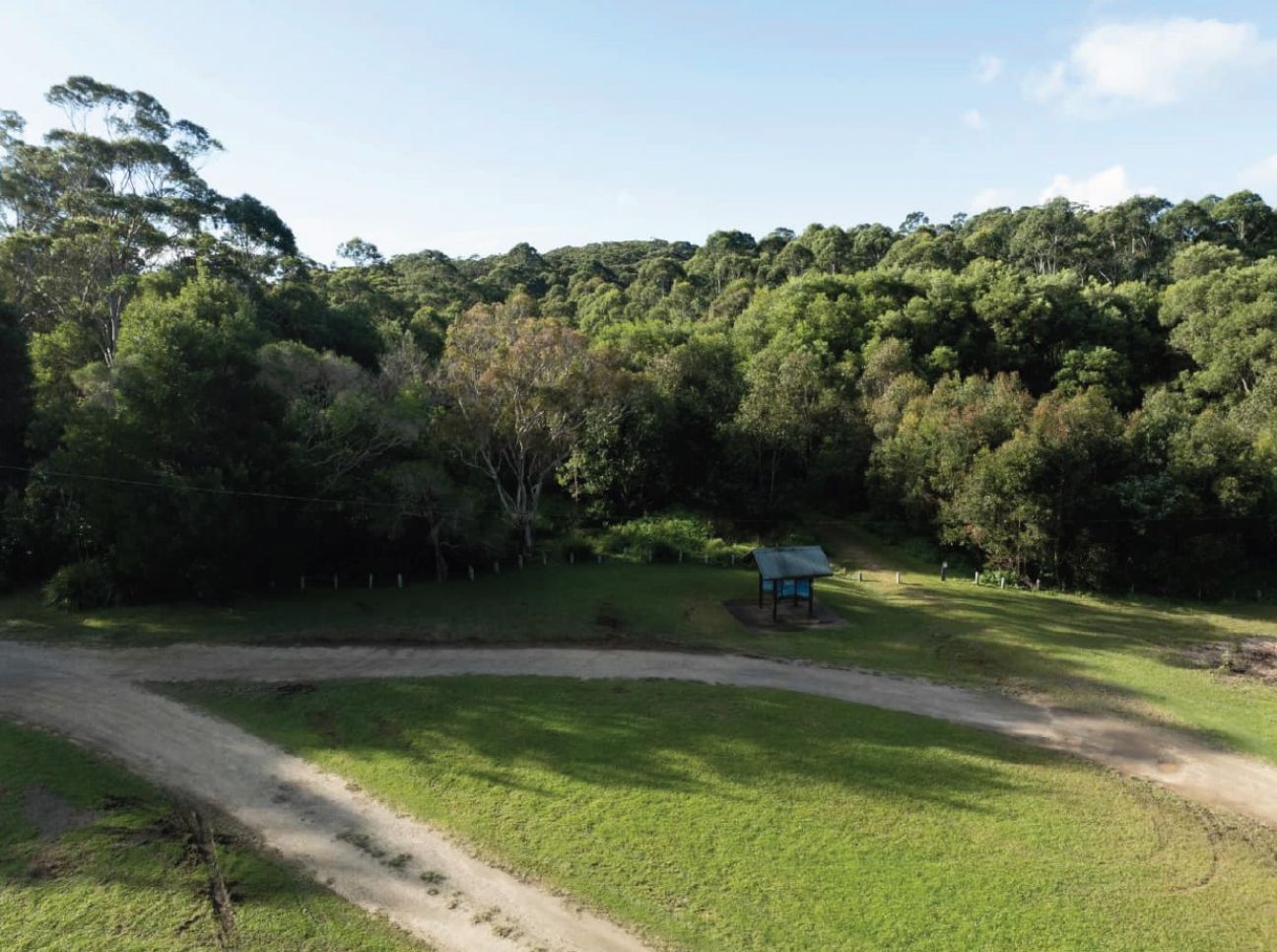 Photo of the Settlement picnic area as it is now—a large grassy area bounded by forest, with a timber information shelter and the start of a walking track