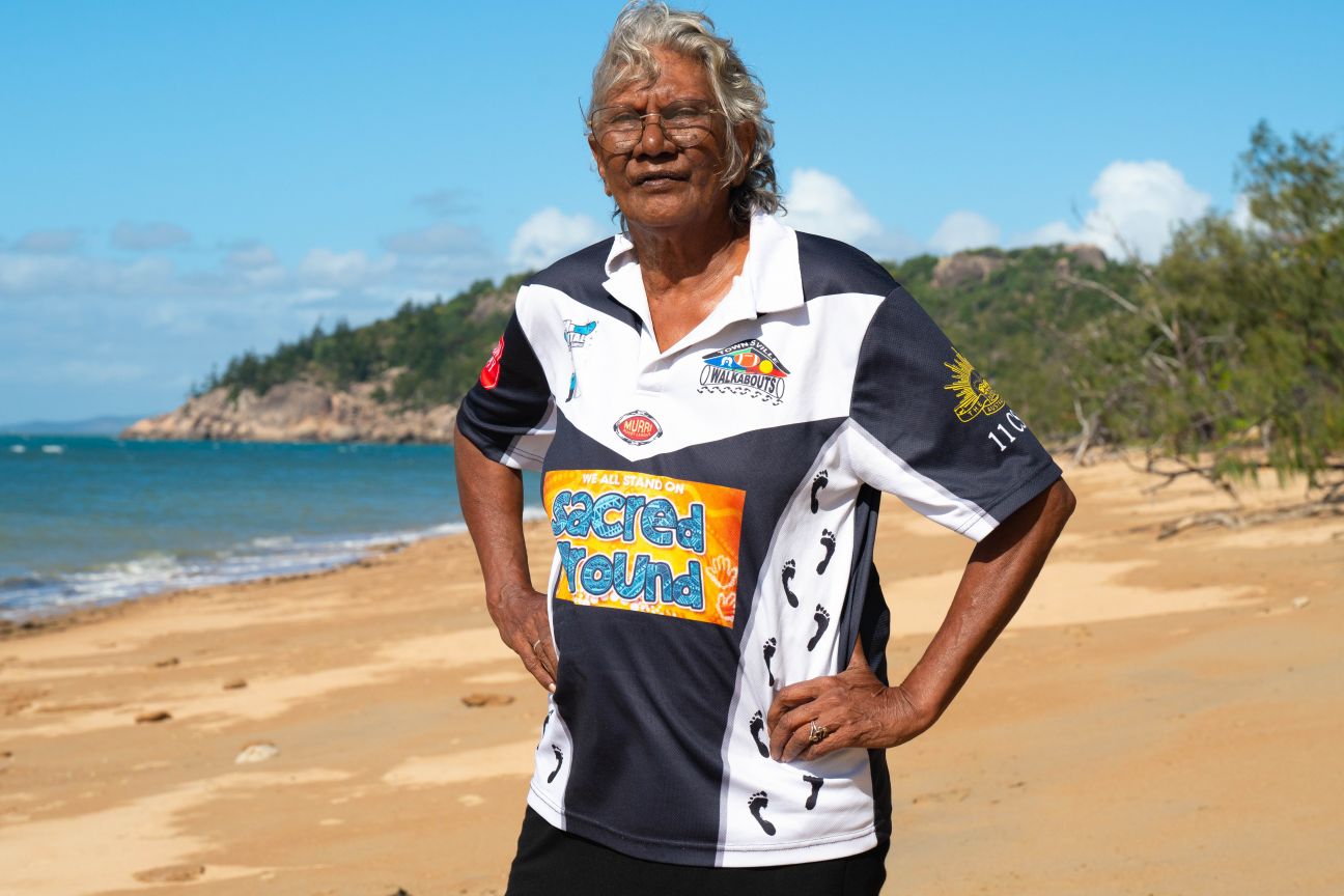Wulgurukaba female elder standing with hands on hips on a beach with the island headland in the background. 