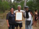 Three Wulgurukaba people smiling standing around a large, framed document with bushland in background.