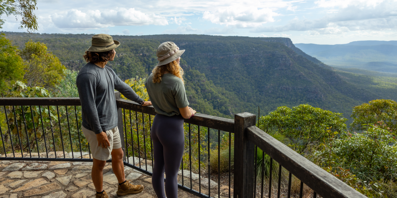 Photo of two people stand at timber fence at lookout, looking out to forested sandstone cliffs.