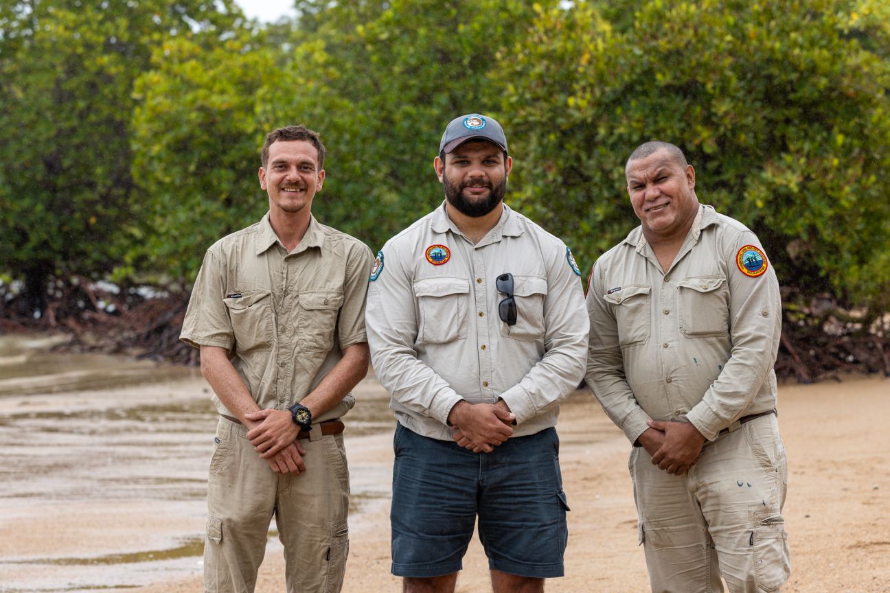 Three Wulgurukaba men wearing Ranger uniforms standing on the beach with mangroves in the background. 