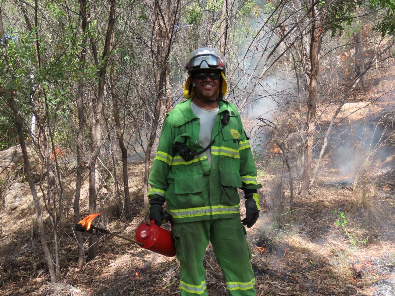 Wulgurukaba man wearing fire uniform holding a drip torch with small fire in the background. 