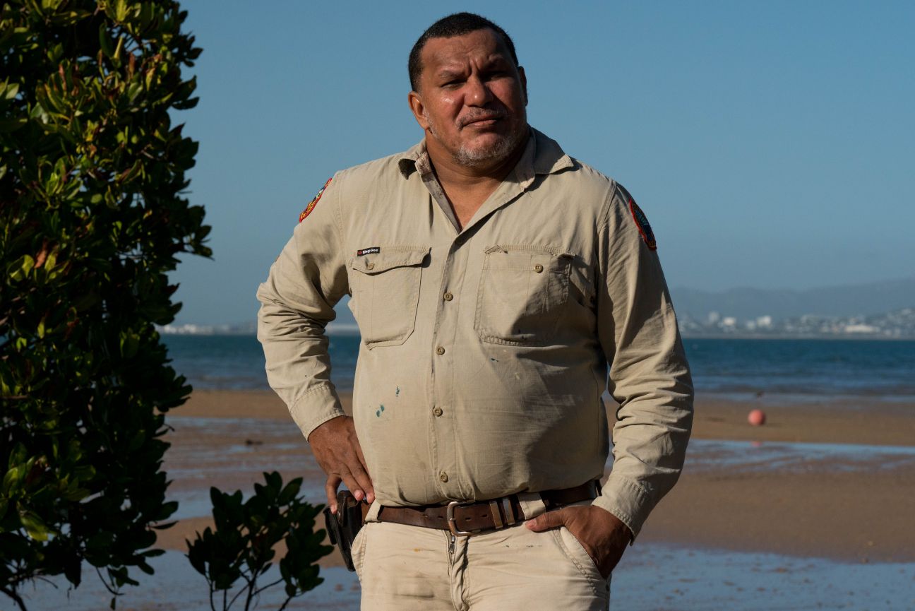 Wulgurukaba man wearing a uniform standing on the beach with mainland in the background. 