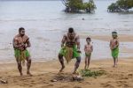 Group of four Wulgurukaba males, two adults and two children, dressed in green lap laps, with white body paint. Standing in front of fire on the beach. One man is using boomerangs as musical instrument.  