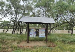 An outdoor information display shelter with a metal roof at Nowranie day-use are. Information shelter is situated in a open grassy area with a tree-lined creek in the background.
