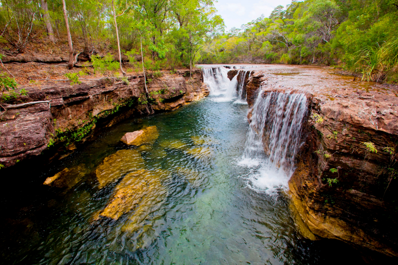 Small waterfalls cascading over large rocky bank into clear water with blue-green tinge