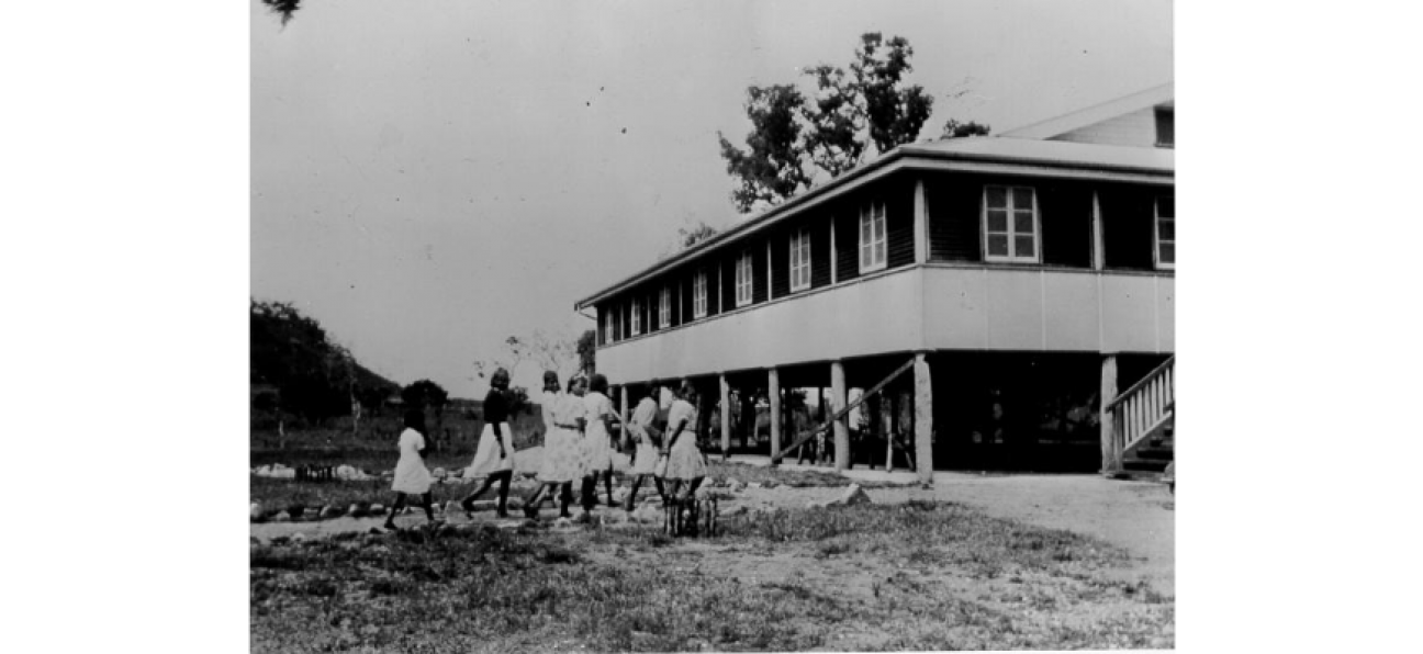 Black and white photo of a large high set Queenslander style building. Group of Aboriginal women wearing white dresses standing in front of building in a cleared area. 