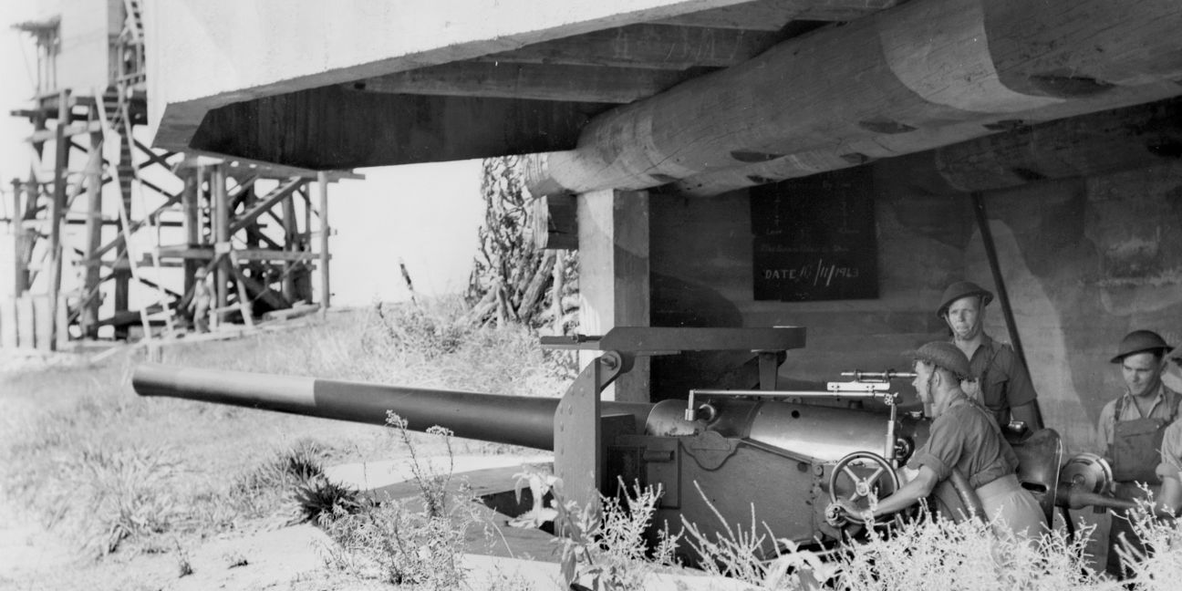 Black and white image of three soldiers standing behind a naval gun within a concrete bunker.