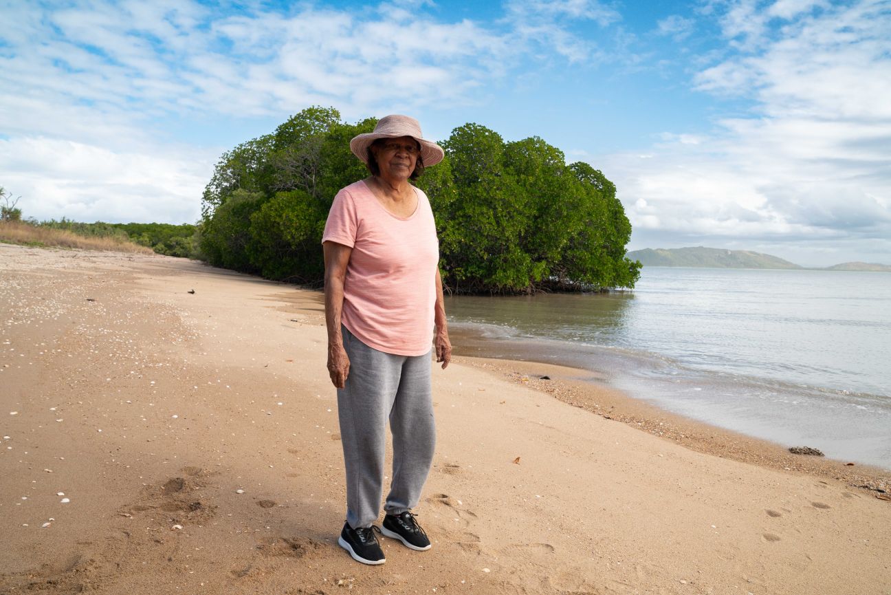 Wulgurukaba female Elder standing on beach with mangroves, ocean and blue sky in background.  