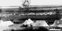 Black and white image of a camp with lots of white tents and soldiers standing in formation.