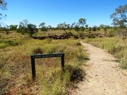 A dirt walking track leading through grassy terrain with scattered trees under a clear blue sky. A green wooden sign labelled  'Little Nowranie Cave