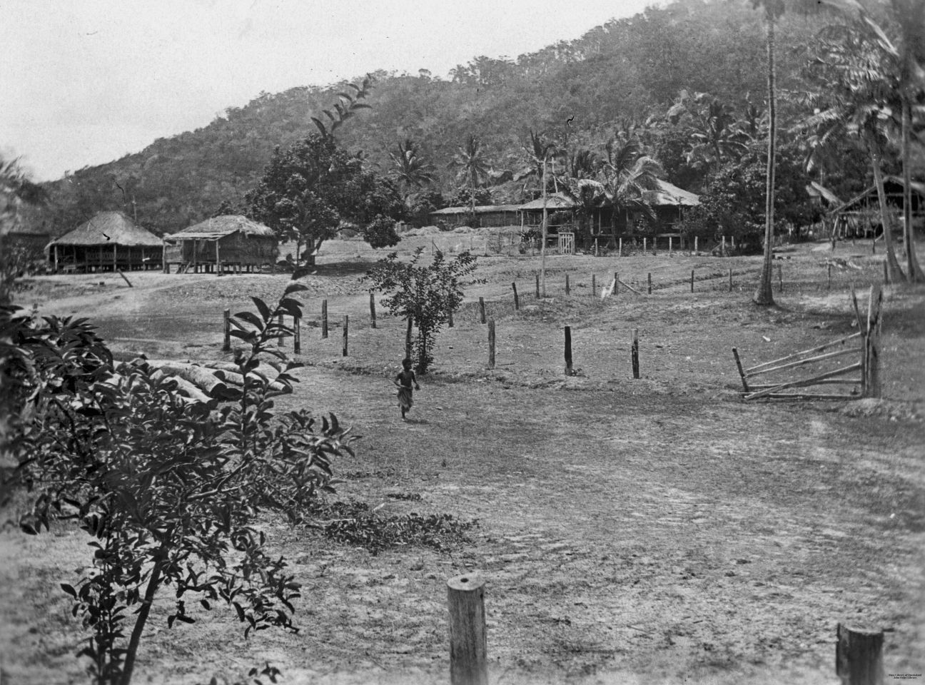 Black and white photo of a rural property with three simple buildings, fence line and some trees. Cleared flat area with forested hills in background.